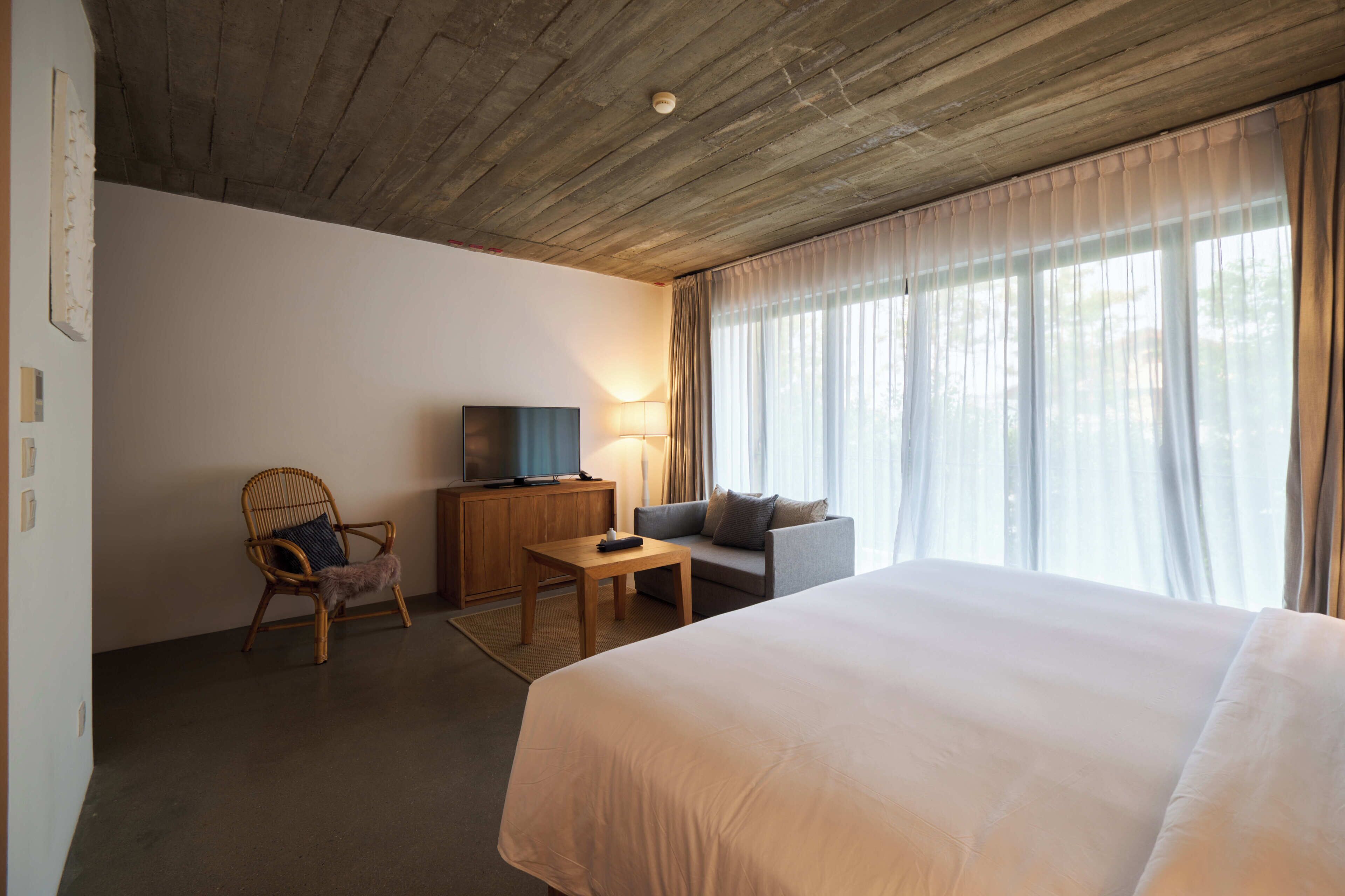 A hotel room with a large bed, a wooden ceiling, a television on a wooden cabinet, a chair, a sofa, and a window with curtains.