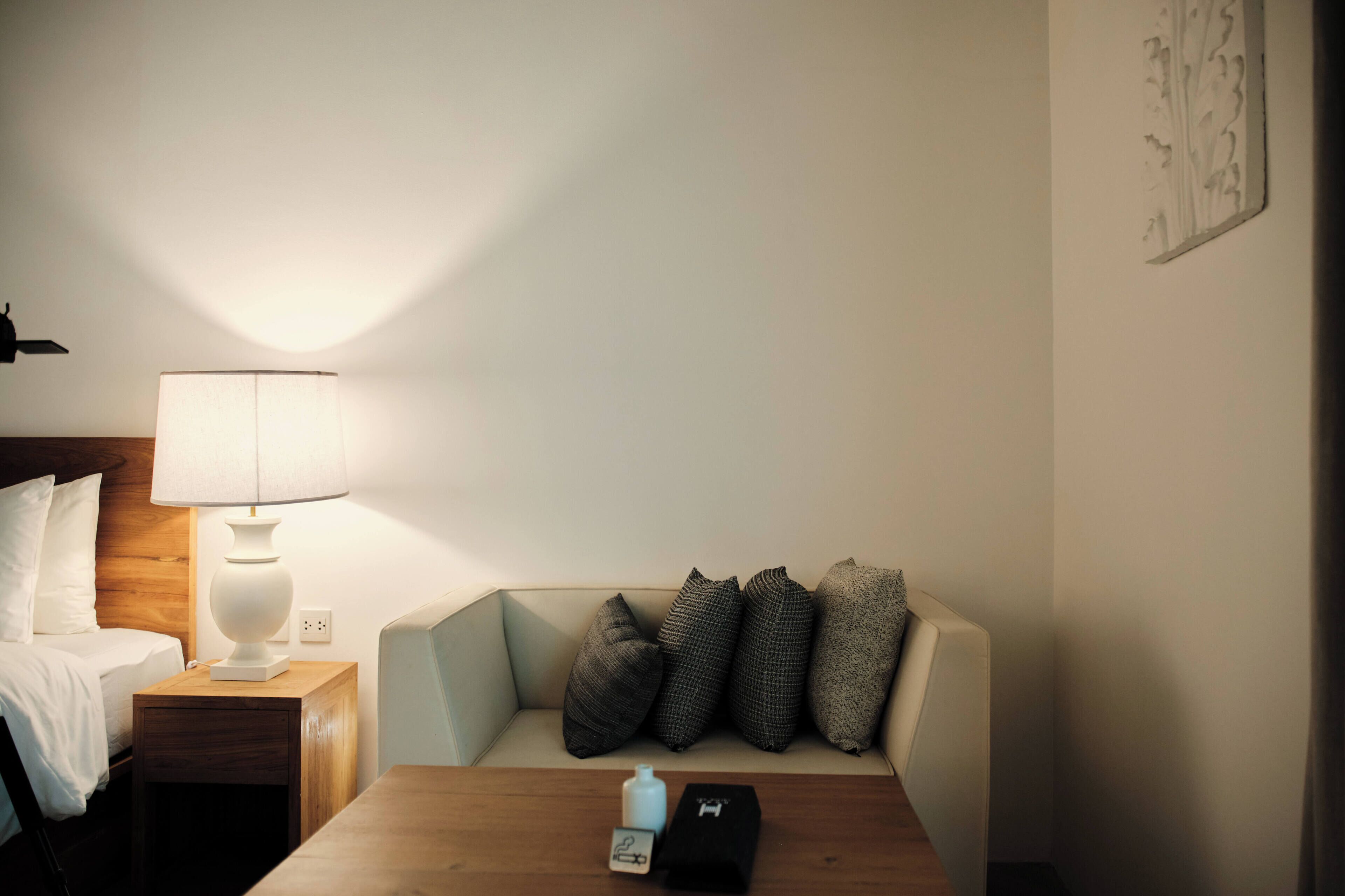 Habitación de hotel con cama, mesita de noche con lámpara, sofá con cojines y una pequeña mesa con una botella y una caja.