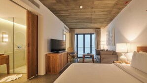 Habitación de hotel con una cama grande, televisión, cómoda de madera, silla, mesita y balcón con vistas.