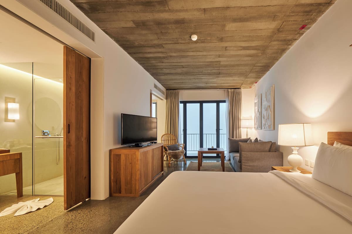 Ein Hotelzimmer mit einem großen Bett, einem Fernseher, einer Holzkommode, einem Sessel, einem kleinen Tisch und einem Balkon mit Blick.