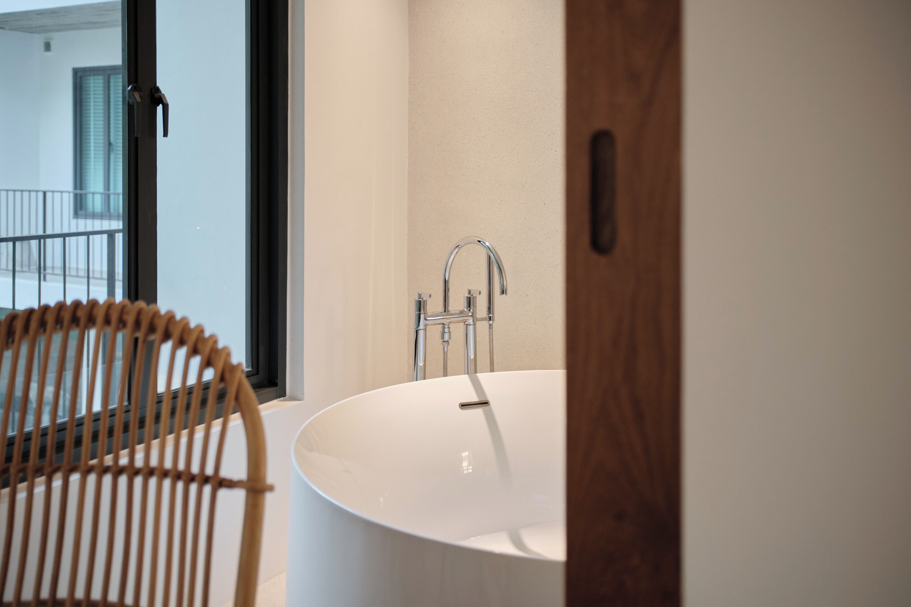 A modern bathroom with a large white bathtub, a wooden chair, and a window with a view of a balcony.