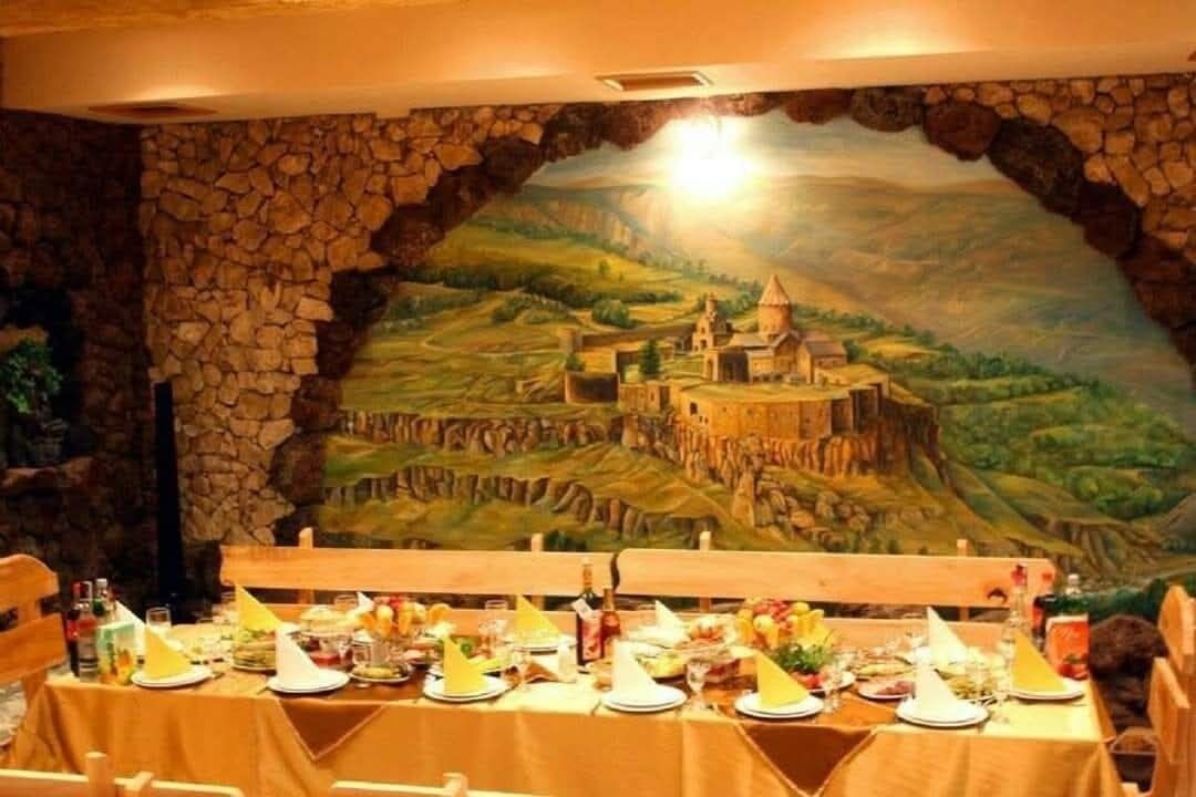 A dining area with a table set for a meal, a stone wall backdrop, and a mural of a landscape with a castle.