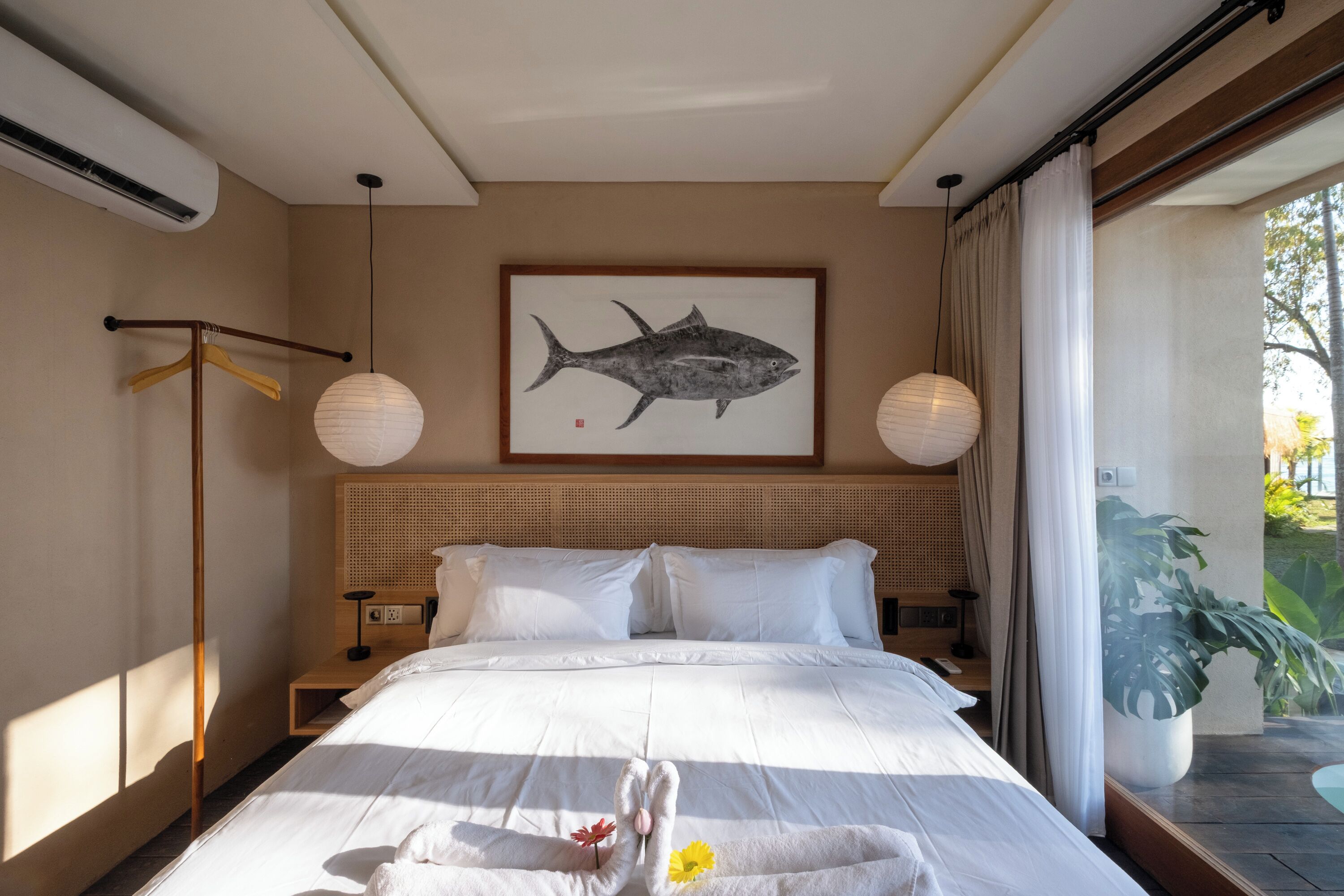 A hotel room with a bed, a wall-mounted artwork of a fish, a bedside lamp, and a view of the outdoors.