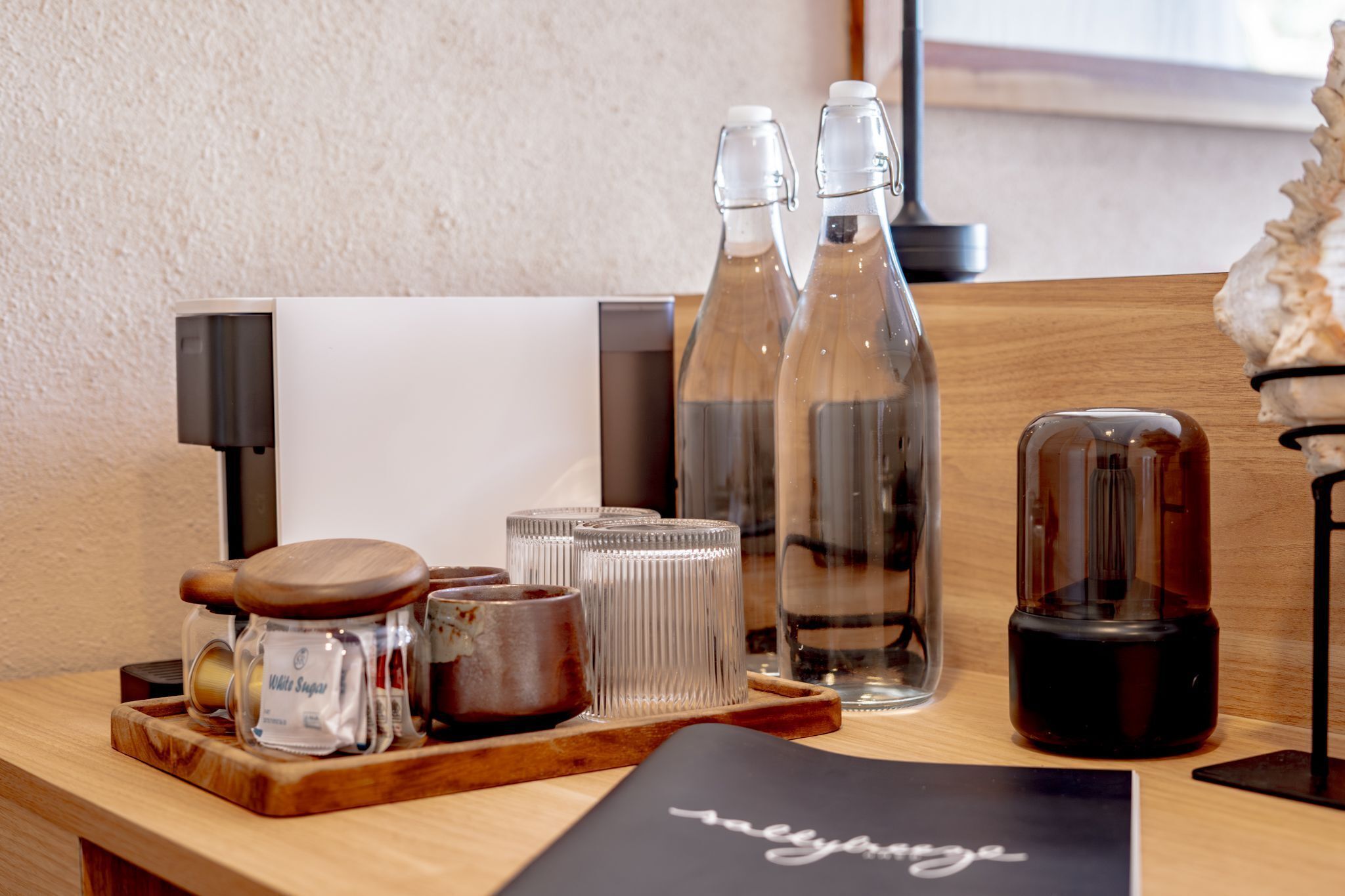 A wooden table with a tray holding a jar of white sugar, two clear bottles, a glass container, and a black object on a black mat.