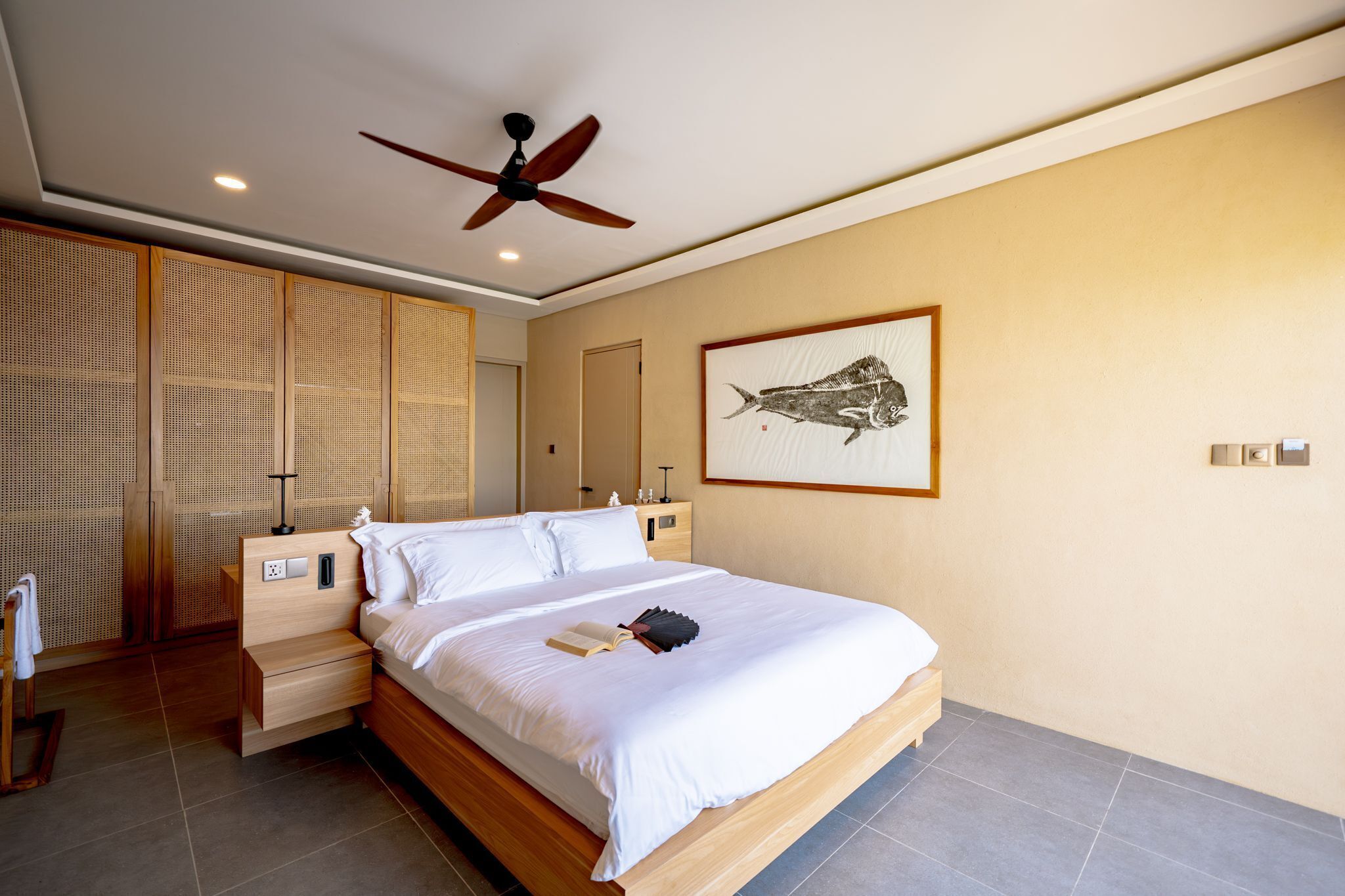 A modern hotel room with a large bed, a ceiling fan, a wall-mounted artwork, and a wooden nightstand.