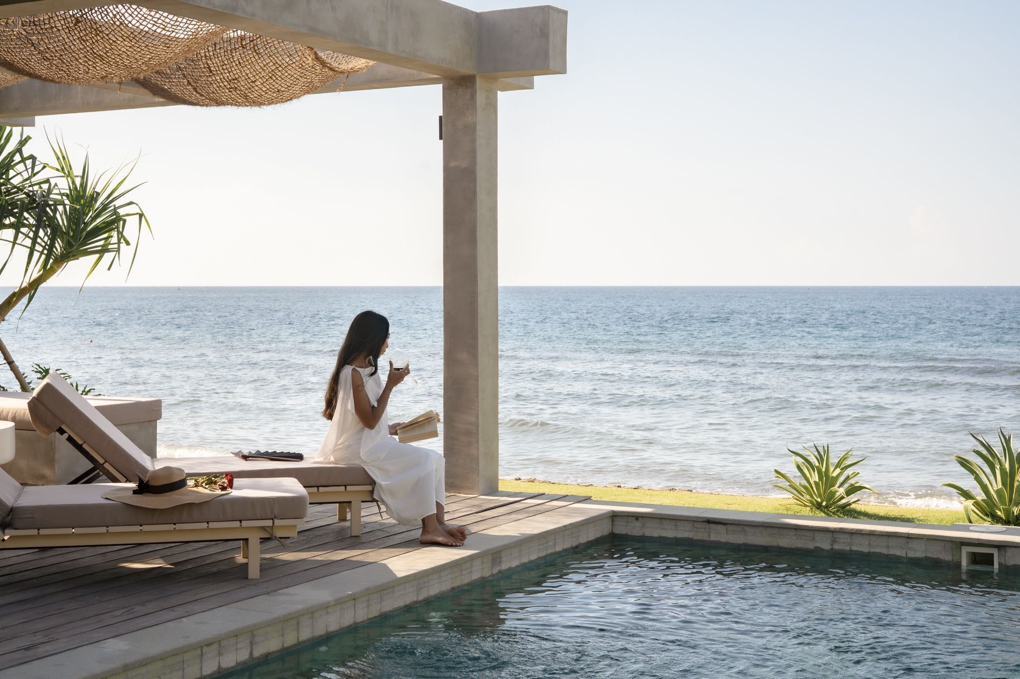 Eine Frau sitzt an einem Pool mit Blick auf das Meer, einem Liegestuhl und einer Betonkonstruktion.