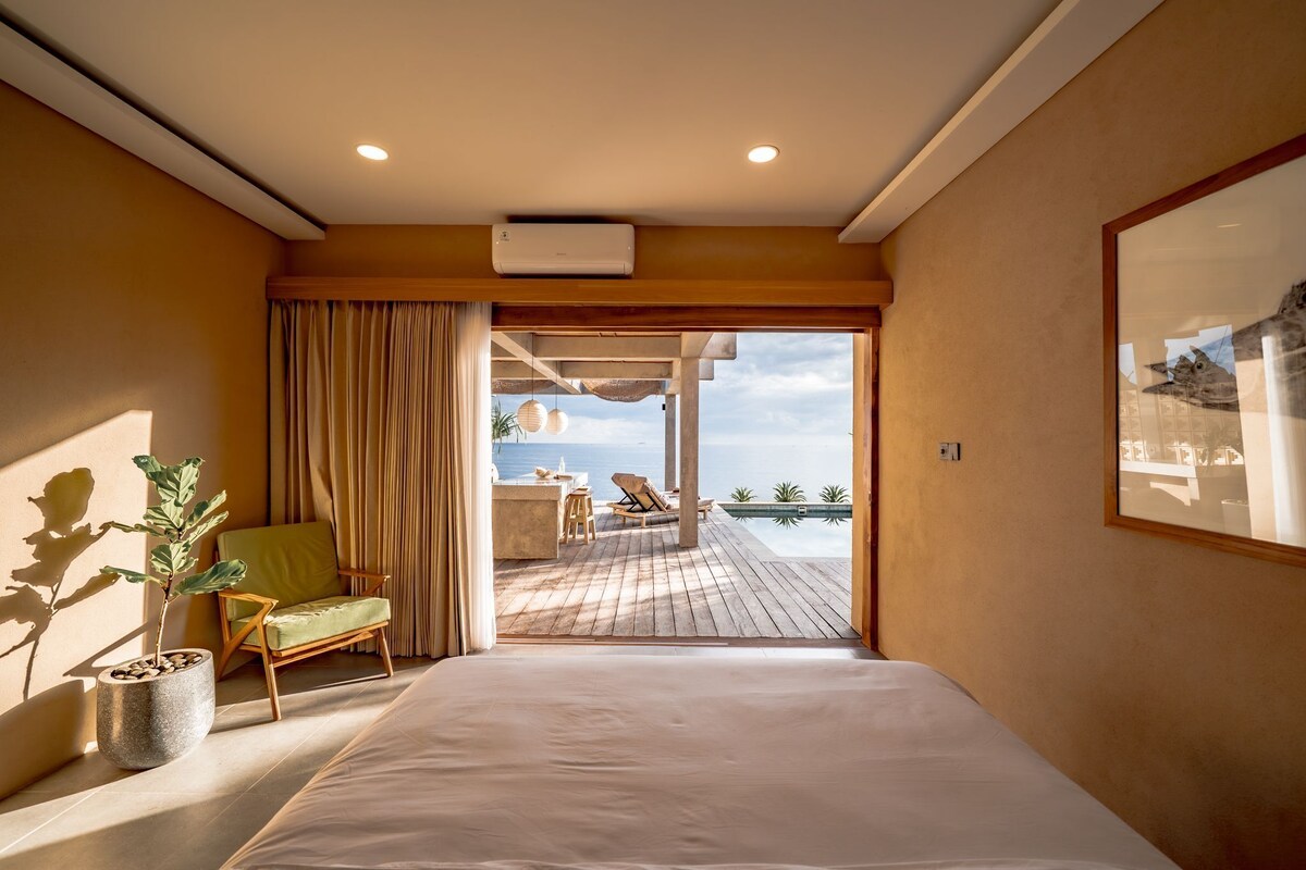 Habitación de hotel con una cama, una silla, una planta en maceta y vista al océano.