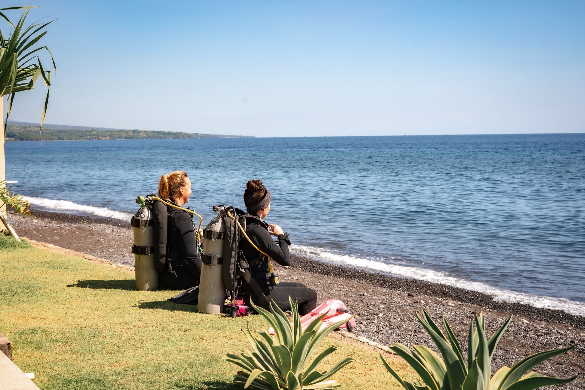 Two individuals seated on a grassy area by a body of water, with scuba diving equipment and a life jacket nearby.