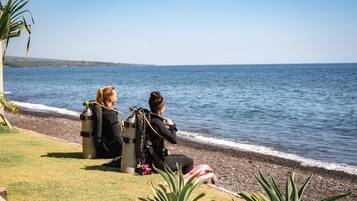 Two individuals seated on a grassy area by a body of water, with scuba diving equipment and a life jacket nearby.