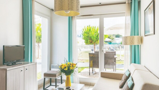 A modern living room with a sofa, a TV, a coffee table with books and a vase of flowers, and a balcony with outdoor furniture. - Comfortable Duplex Retreat for Family (Saint-Palais-sur-Mer)