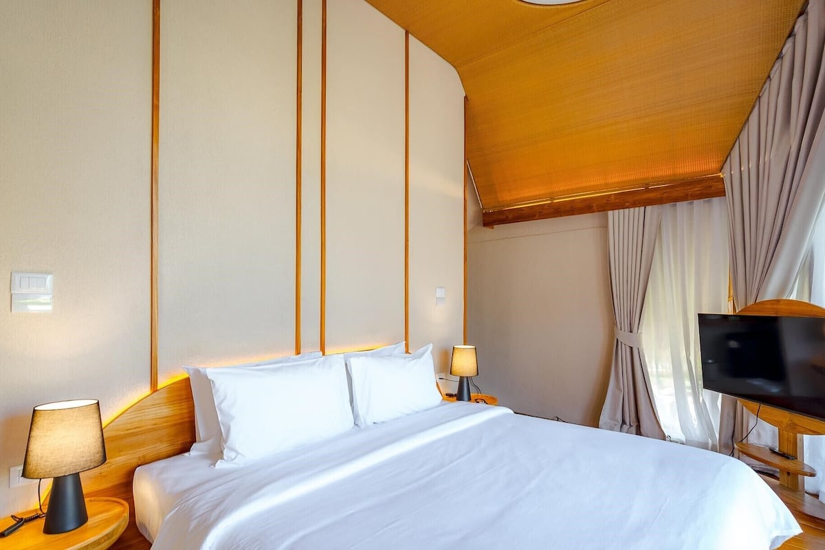 A hotel room with a bed, two bedside tables with lamps, a flat-screen TV, and a large window with curtains.