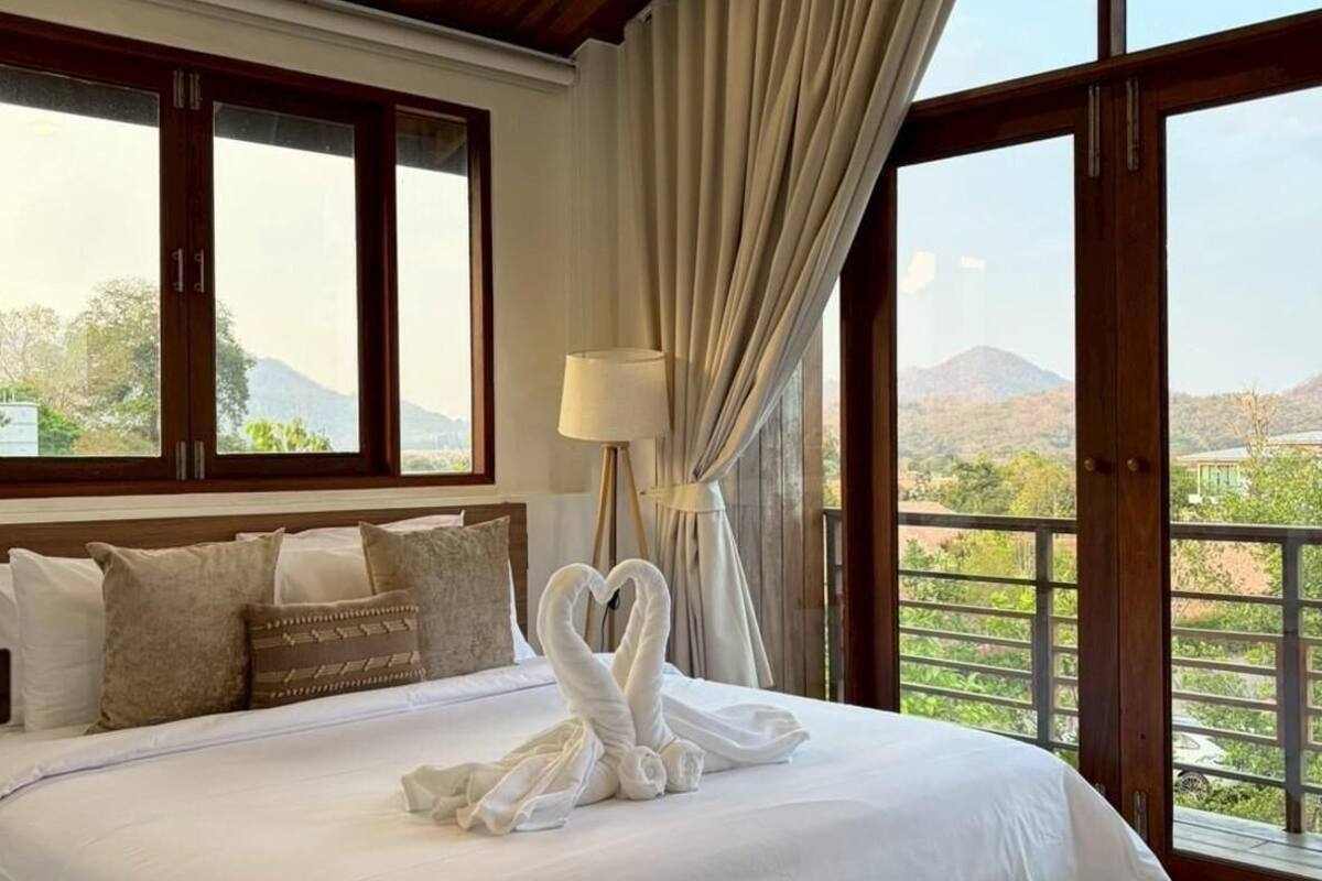 A large bed with white linens, a wooden ceiling, and a view of greenery through large windows.