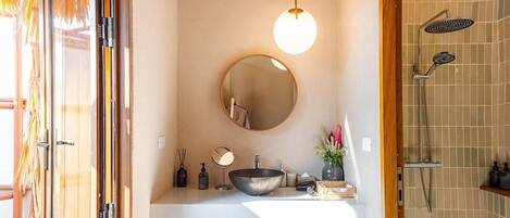 A bathroom with a wooden vanity, a round mirror, a hanging light, a shower with a rain showerhead, and a tiled wall.