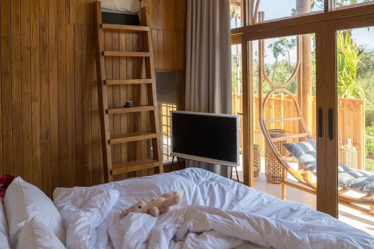 A bedroom with a bed, a TV, a wooden ladder, and a view of the outdoors.