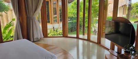 A room with a bed, a TV, and large windows offering a view of greenery and a building.