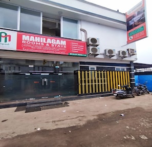 A building with a sign for 'MAMILAGAM ROOMS & STAYS' advertising amenities like A/C rooms, 24-hour water, WiFi, and more.
