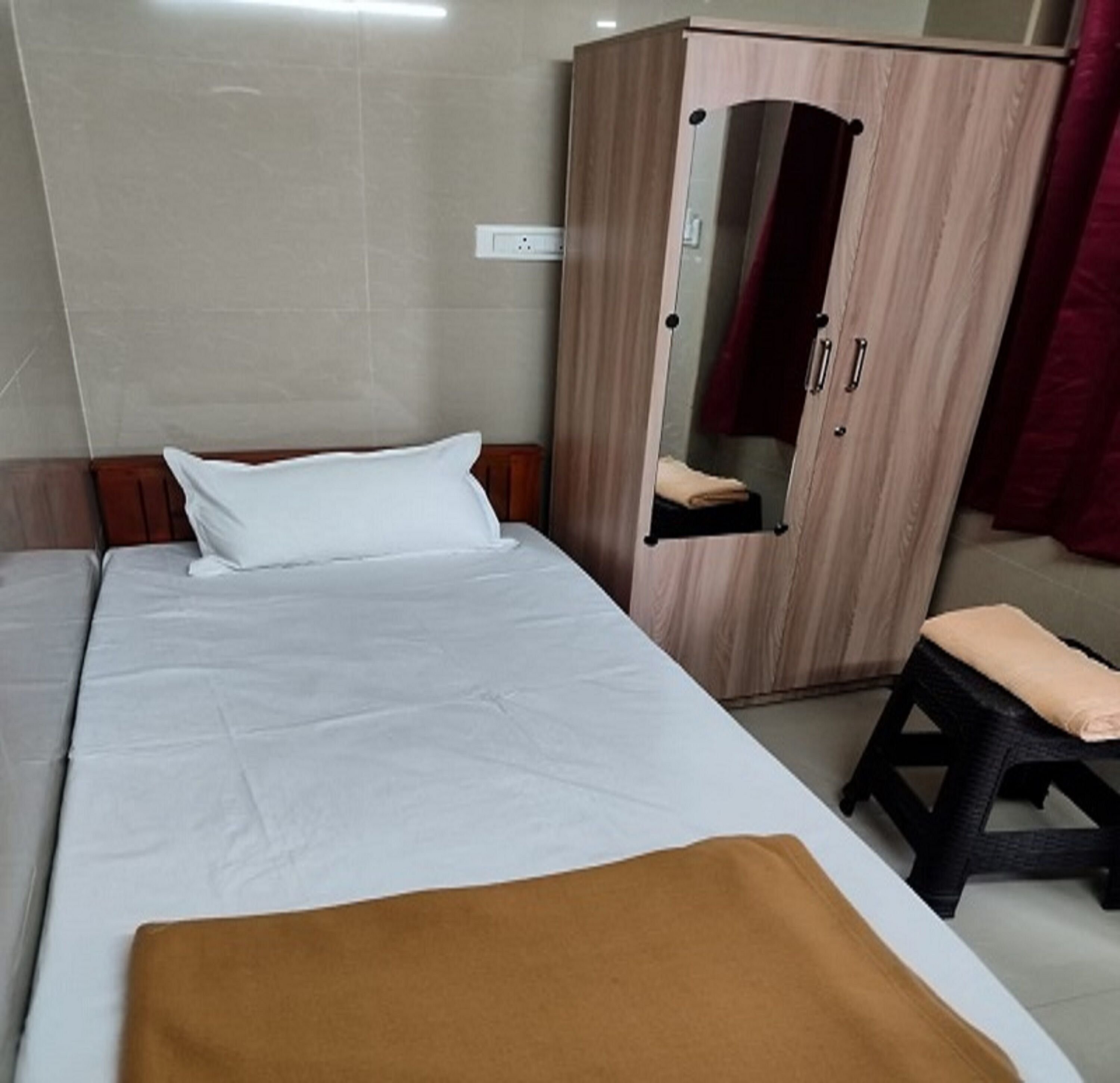 A single bed with a white mattress and a brown blanket, a wooden wardrobe with a mirror, and a small stool.