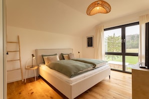 A bedroom with a bed, a ladder, a bedside table, a lamp, and a large window with a view of greenery.