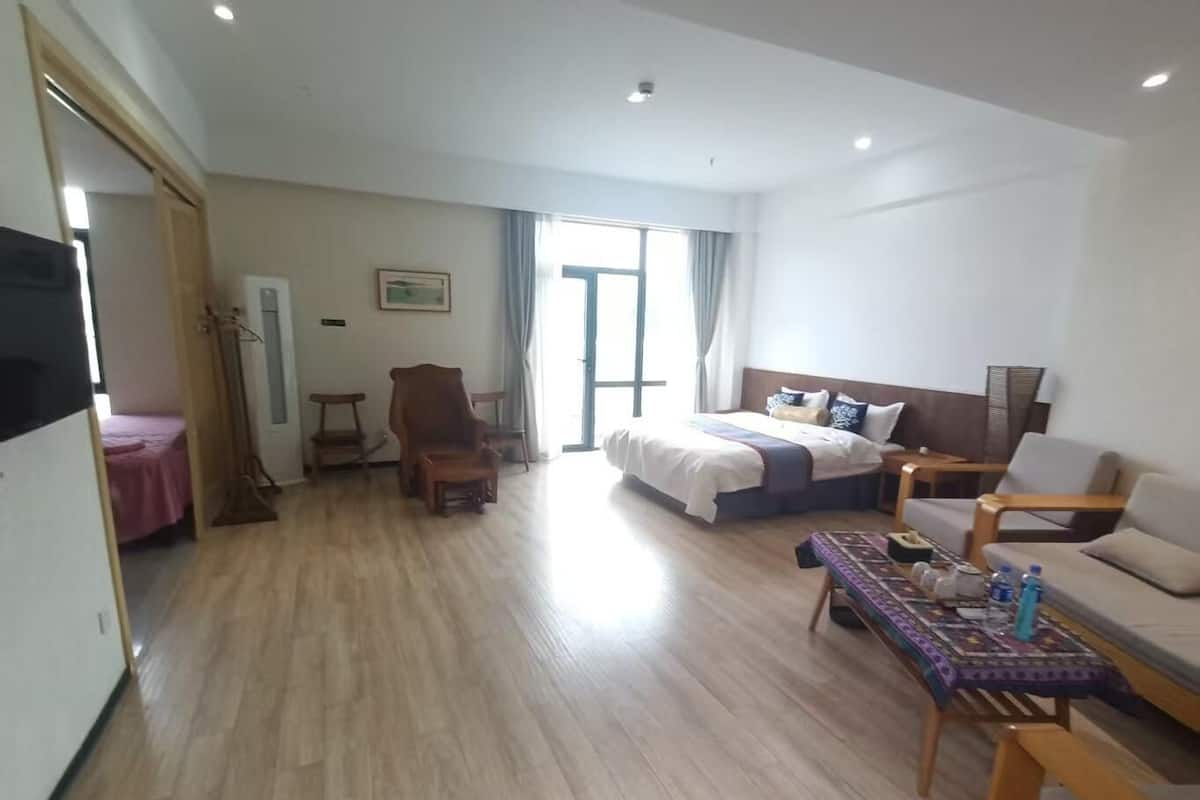 A hotel room with a bed, a sofa, a table, and a chair. There is a window with curtains, a door, and a wall-mounted picture.
