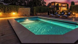Una piscina rettangolare con bordo piastrellato, circondata da sdraio e un edificio ben illuminato sullo sfondo.