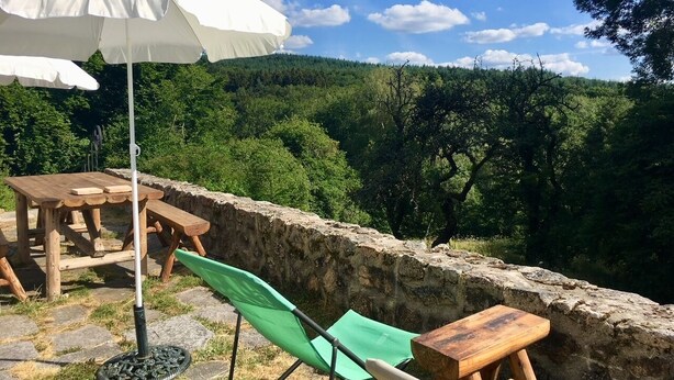 Une terrasse avec une table et des chaises en bois, un parasol blanc et un mur en pierre.