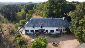 Une maison blanche avec un toit foncé, entourée d’arbres et bordée d’un chemin de terre.