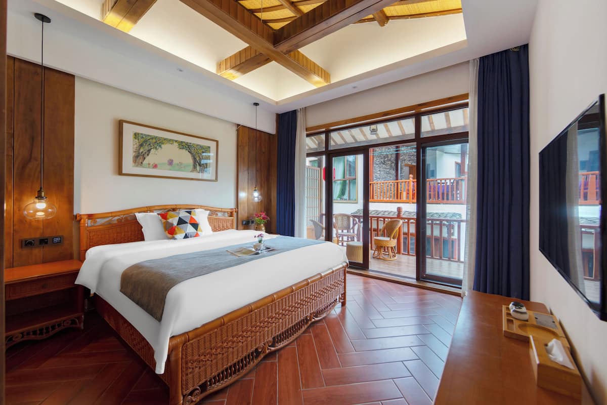 Habitación de hotel con una cama grande, suelo de madera, balcón con mobiliario exterior y un ventanal amplio.
