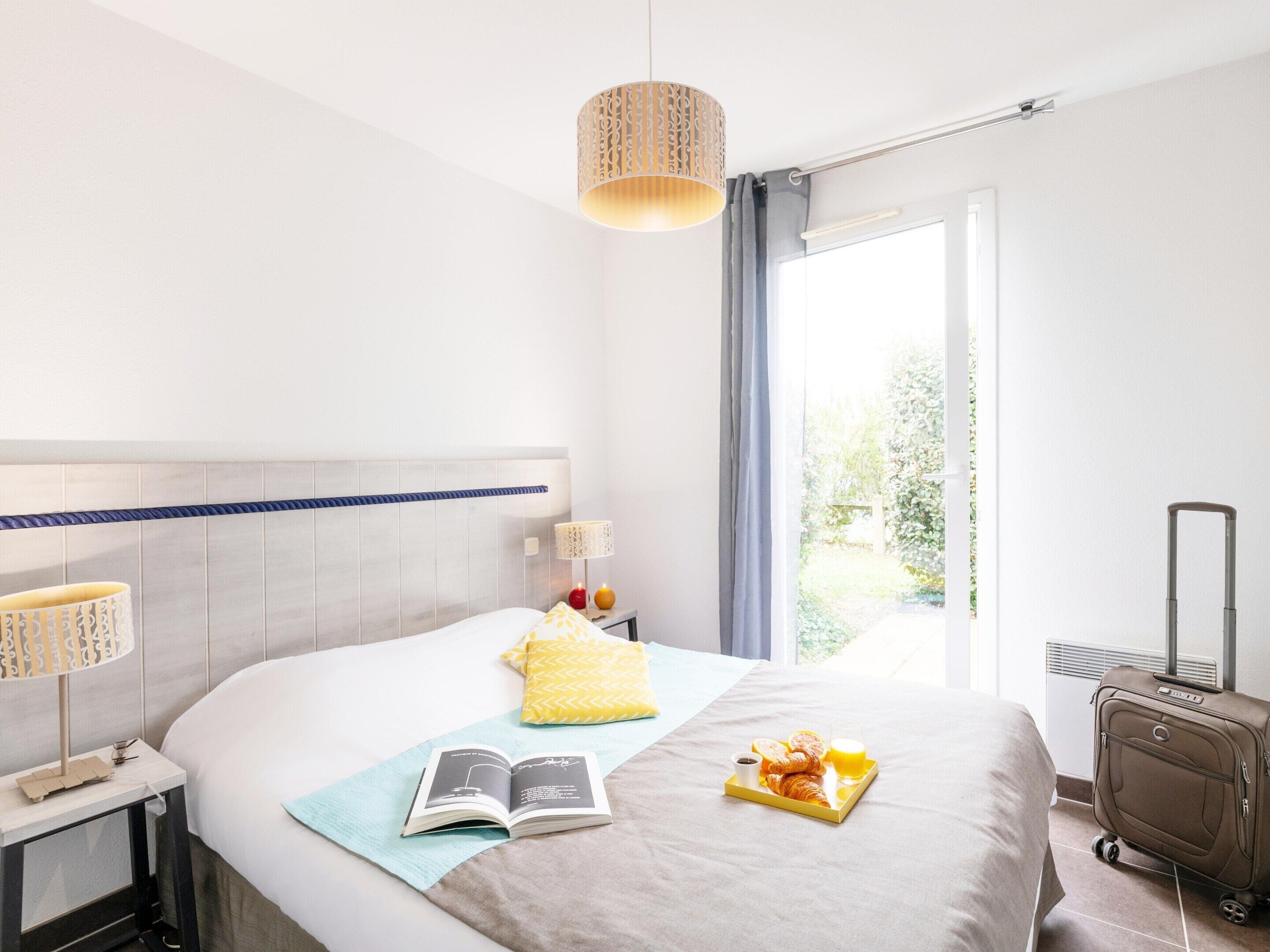 Ein Hotelzimmer mit einem Bett, einem Nachttisch mit Lampe, einem Koffer und einem Fenster mit Vorhängen.
