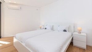 Un dormitorio moderno y minimalista con dos camas individuales, ropa de cama blanca, mesitas de noche con lámparas y suelo de madera.