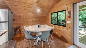 A wooden cabin interior with a round table, chairs, a refrigerator, and a window overlooking greenery. - Bear Ridge BaseCamp at Buffalo Creek | Pet-Friendly Lake Lure Cabin with Views, Firepit & Game Room (Lake Lure)