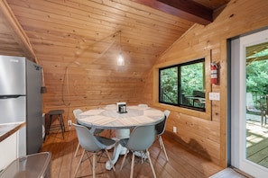 Een houten hut met een ronde tafel, stoelen, een koelkast en een raam met uitzicht op groen.