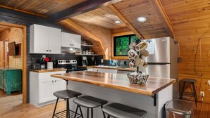 A modern kitchen with wooden beams, a large island with bar stools, white cabinets, and a stainless steel refrigerator. - Bear Ridge BaseCamp at Buffalo Creek | Pet-Friendly Lake Lure Cabin with Views, Firepit & Game Room (Lake Lure)