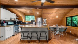 A modern kitchen with wooden walls, a central island with bar stools, a refrigerator, and a dining area with a table and chairs. - Bear Ridge BaseCamp at Buffalo Creek | Pet-Friendly Lake Lure Cabin with Views, Firepit & Game Room (Lake Lure)