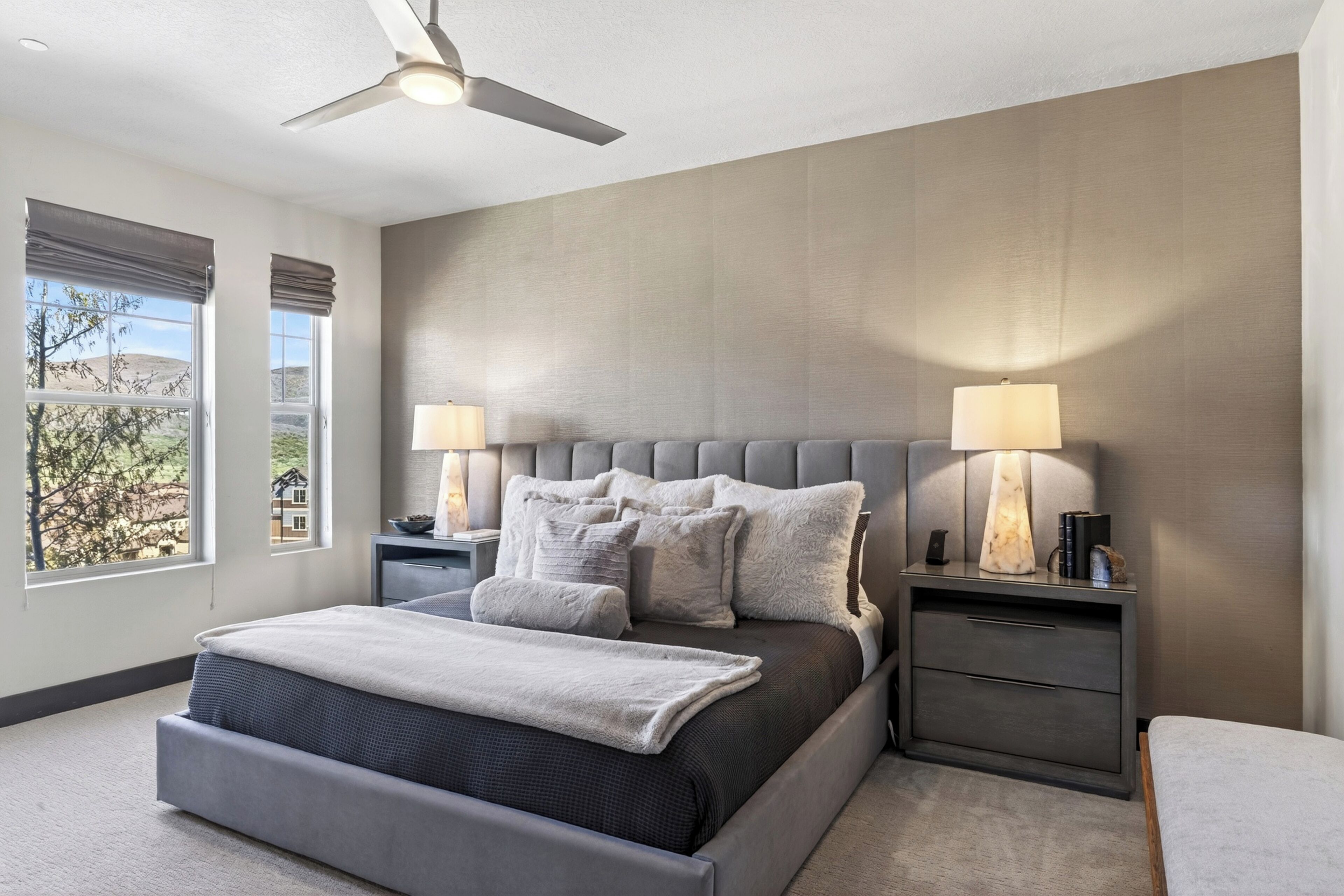 Une chambre à coucher avec un lit, deux tables de chevet, un ventilateur de plafond et de grandes fenêtres.