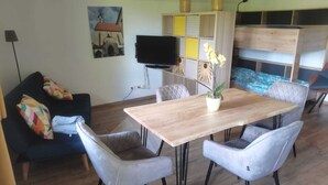 Una sala de estar con una mesa de comedor de madera, sillas tapizadas en gris, un sofá azul con cojines de colores, un televisor de pantalla plana y un panel decorativo en la pared.