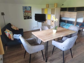 Una sala de estar con una mesa de comedor de madera, sillas tapizadas en gris, un sofá azul con cojines de colores, un televisor de pantalla plana y un panel decorativo en la pared.