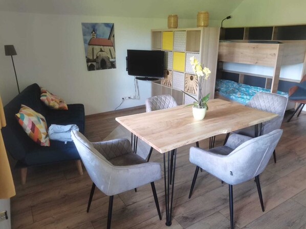 A living room with a wooden dining table, grey upholstered chairs, a blue sofa with colorful pillows, a flat-screen TV, and a decorative wall panel.