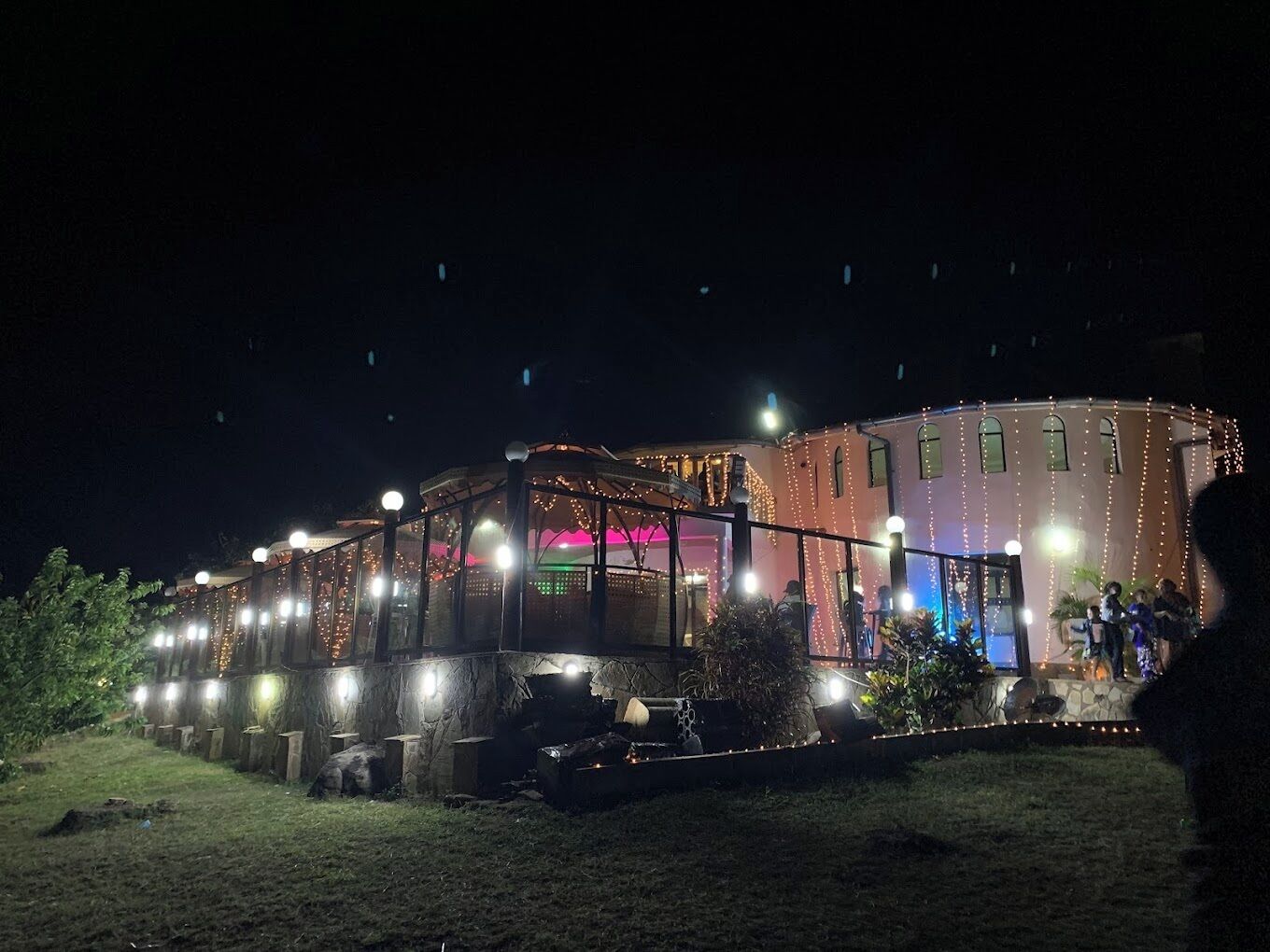Un edificio con un espacio exterior techado, posiblemente un restaurante o bar, con luces colgantes y algunas personas al fondo.