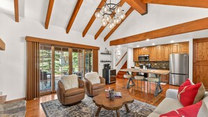 A spacious living area with a large window, wooden ceiling beams, a dining area, and a kitchen with wooden cabinets. - Luxury Tamarack Cottage | Ski-In/Ski-Out | Hot Tub | Walk to Village (Donnelly)