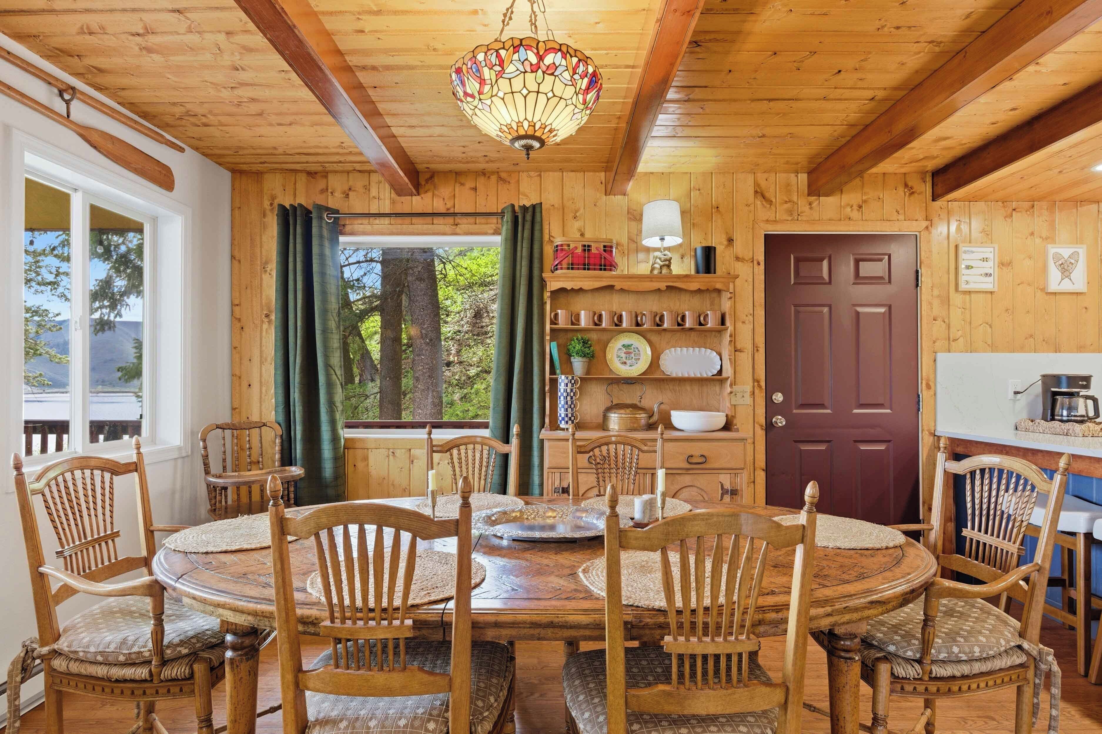 Un interior de madera con una mesa redonda de comedor, sillas, una lámpara de araña y vistas a través de una ventana.