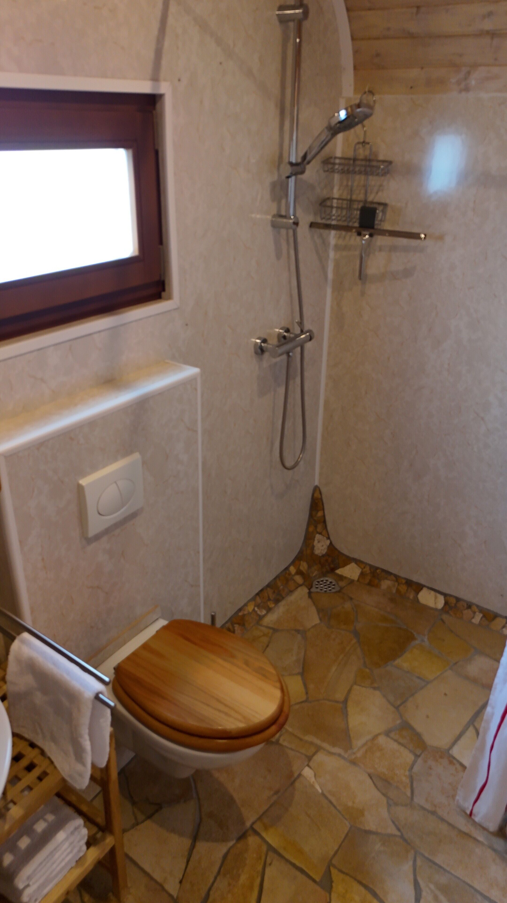 Une salle de bain avec des toilettes en bois, une douche avec un pommeau de douche amovible, un porte-serviettes et une fenêtre.