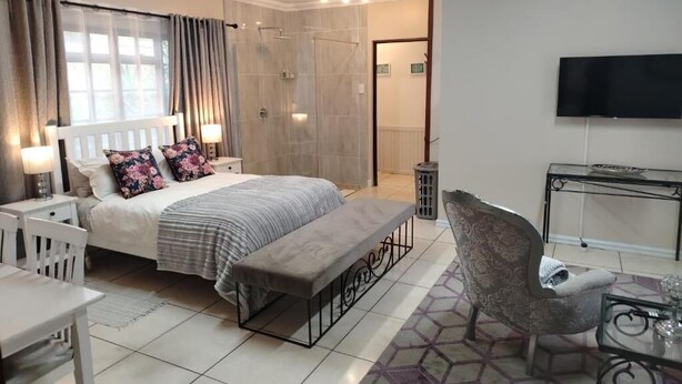 Ein Schlafzimmer mit Bett, Nachttischen, einer Bank, einem Sessel, einem Fernseher und einer Dusche.
