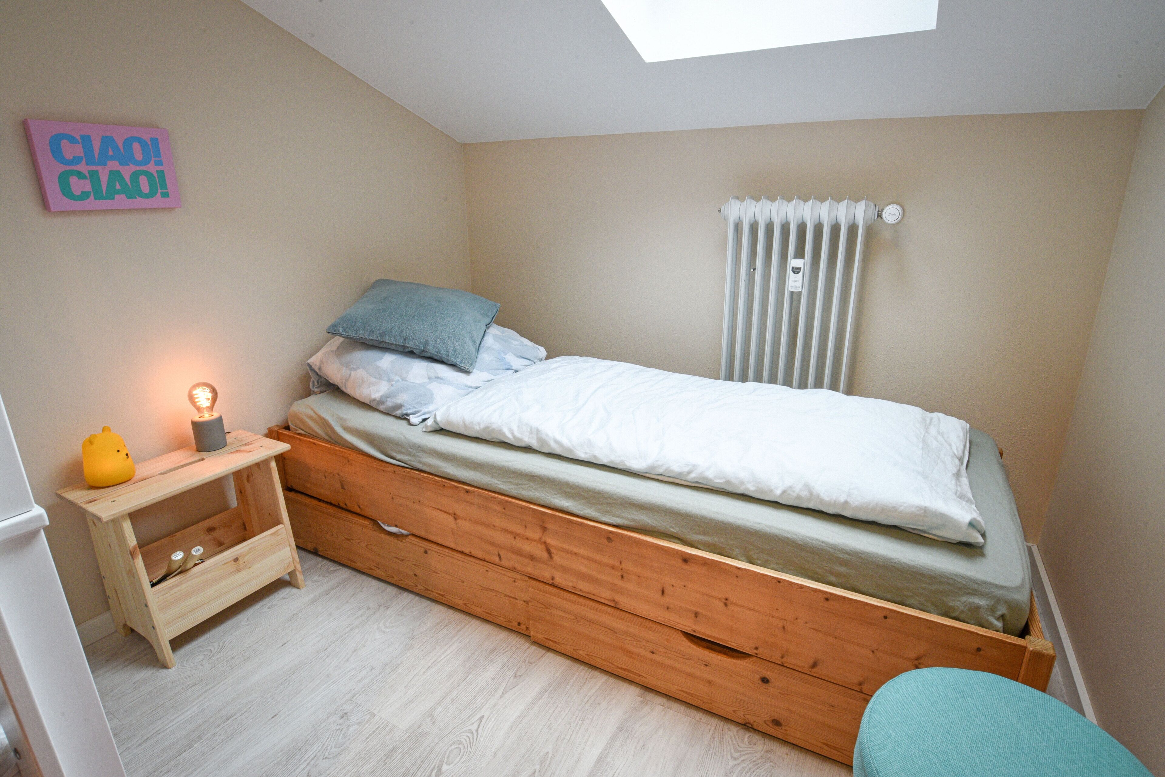 A bedroom with a skylight, a bed with pillows, a bedside table with a lamp, and a radiator.