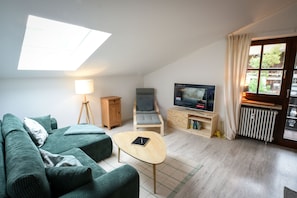 Ein Wohnzimmer mit einer Couch, einem Sessel, einem Tisch, einer Lampe, einem Fernseher und einem Fenster mit Vorhängen.