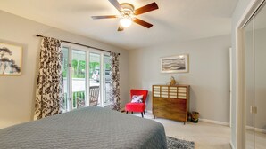 Un dormitorio con cama, ventilador de techo, una silla roja, cómoda de madera y puerta corrediza de vidrio que da a un balcón.