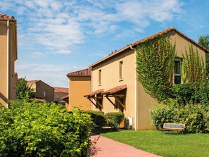 Un edificio residencial con techo de tejas, un porche cubierto y un jardín bien cuidado con un sendero y un banco.