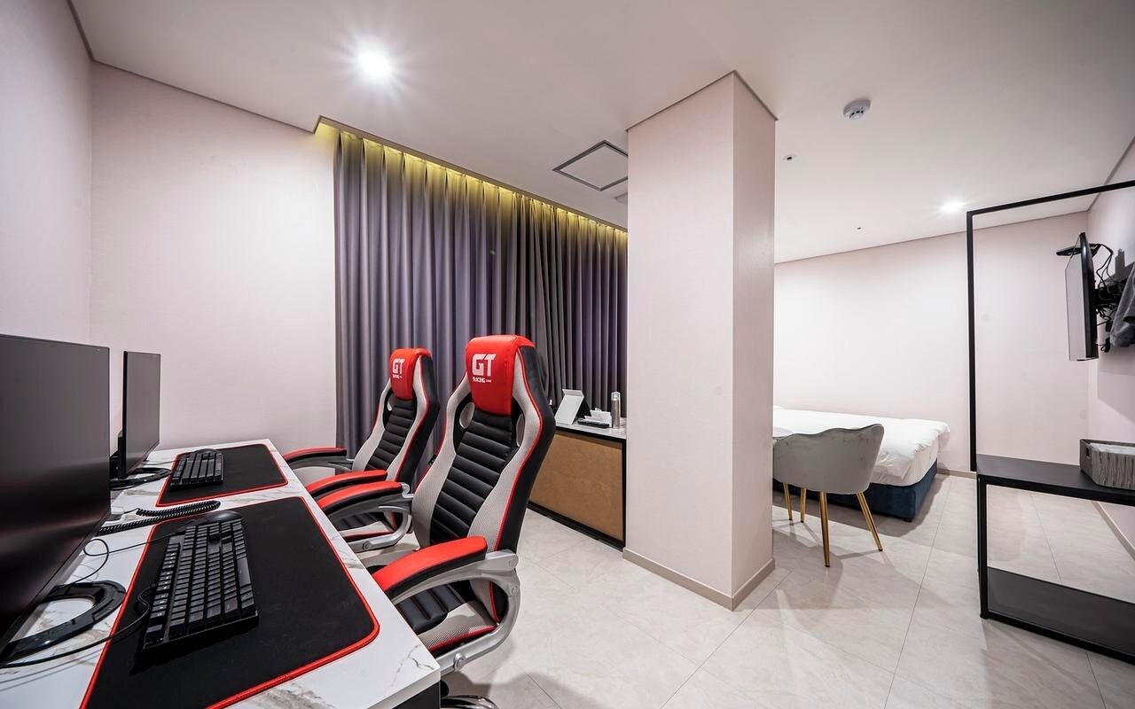 A modern hotel room with a desk, two GT branded gaming chairs, a bed, a small table, and a mirror.