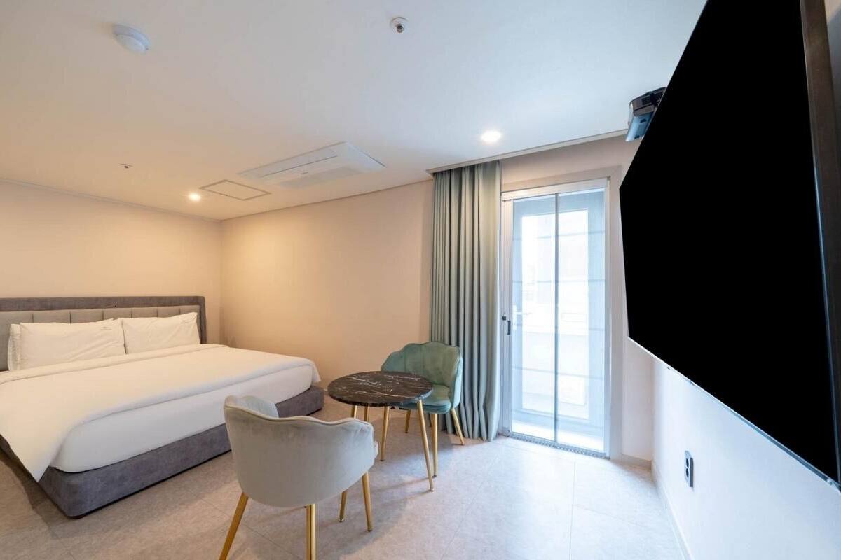 A hotel room with a bed, a TV, a chair, a small table, and a sliding door leading to a balcony.