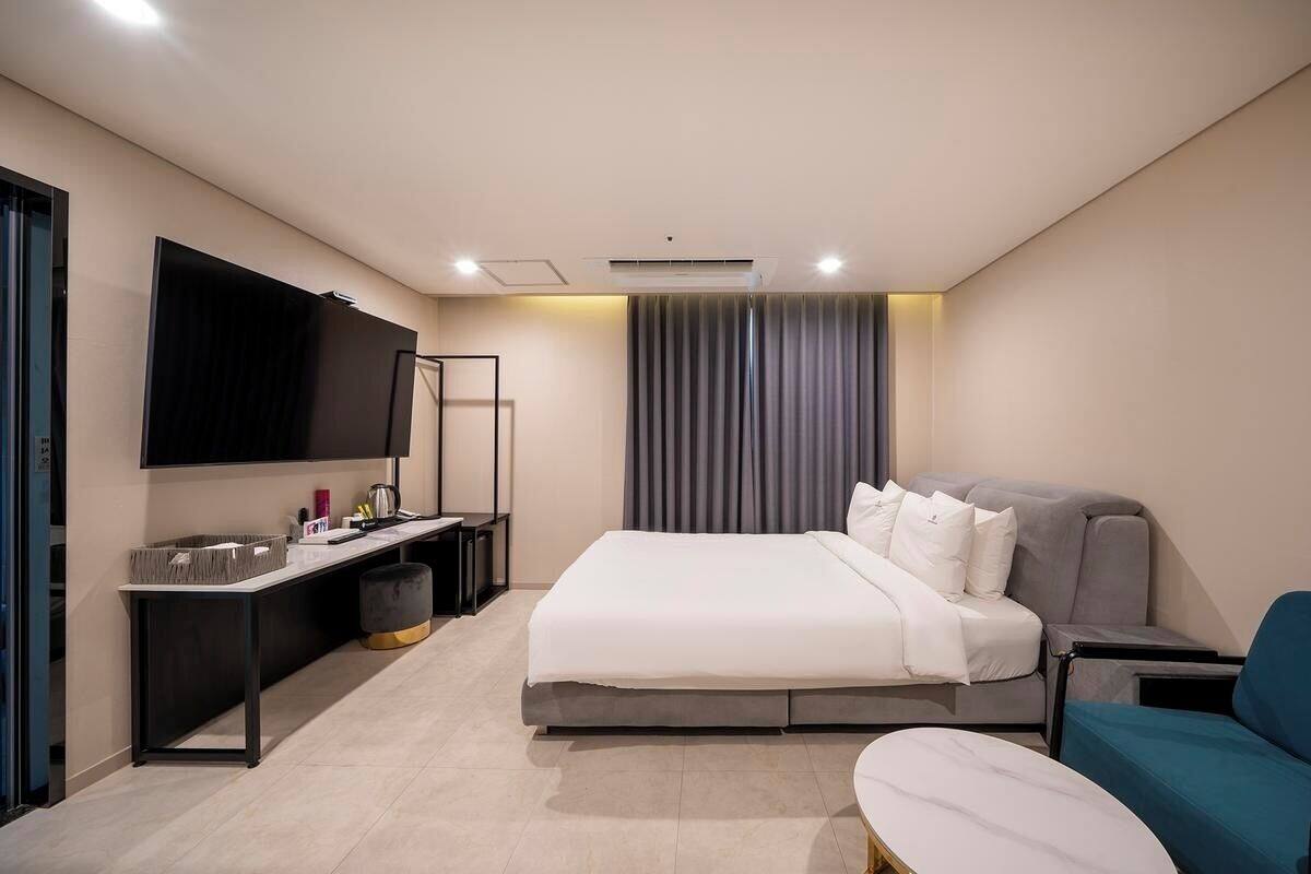 A modern hotel room with a bed, a desk, a TV, and a seating area.