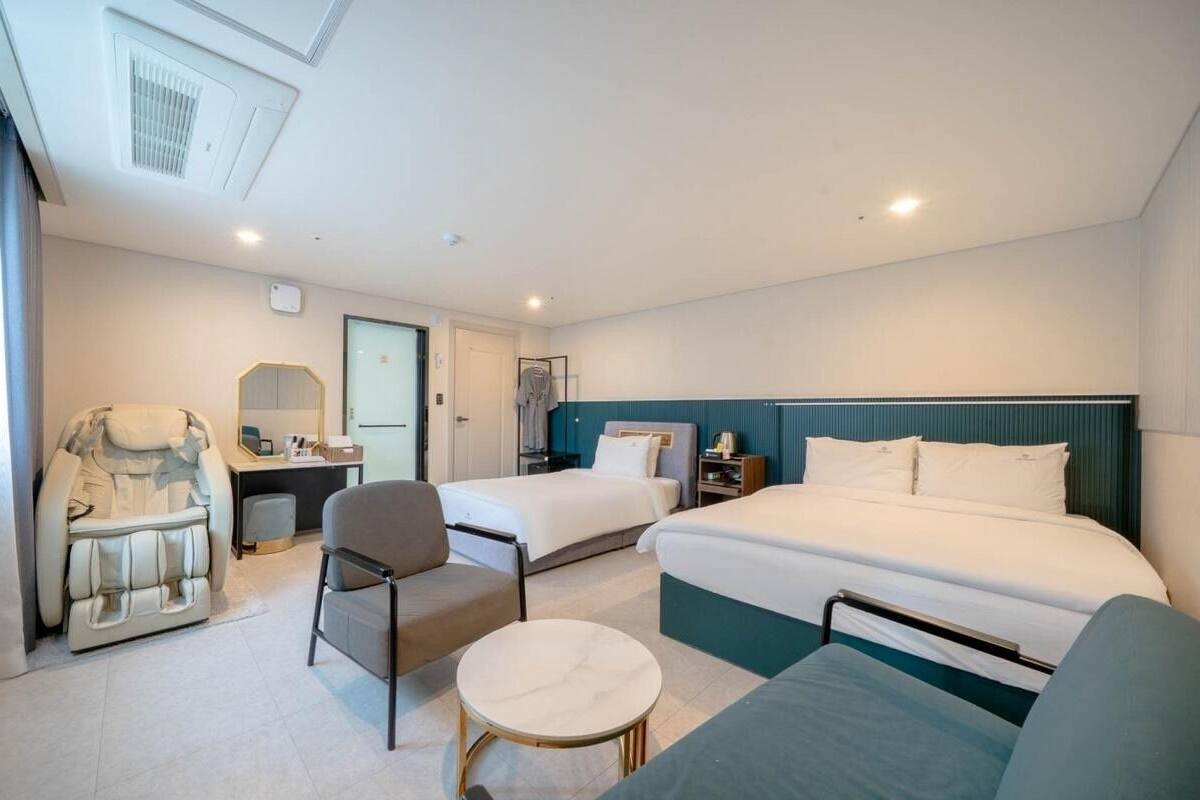 A hotel room with a bed, two chairs, a massage chair, a small table, and a desk with a mirror.